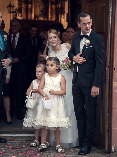 Hochzeitspaar trotzt dem Regen und schaut mit Blumenkinder aus der Kircheneingangstür